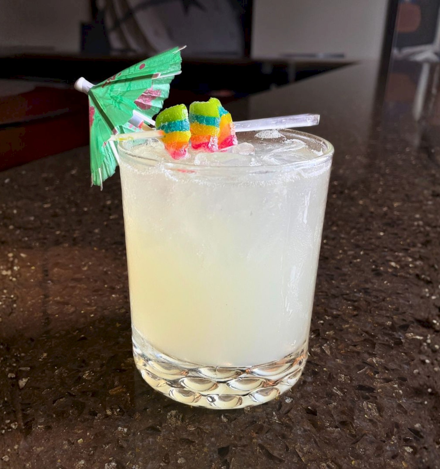 A cocktail in a short glass with ice, garnished with a small umbrella and colorful fruit-shaped candies on a pick.