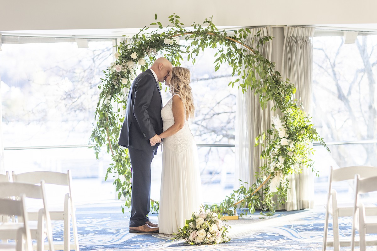 A couple is standing close, holding hands in front of a circular greenery arch, suggesting a wedding setting indoors.