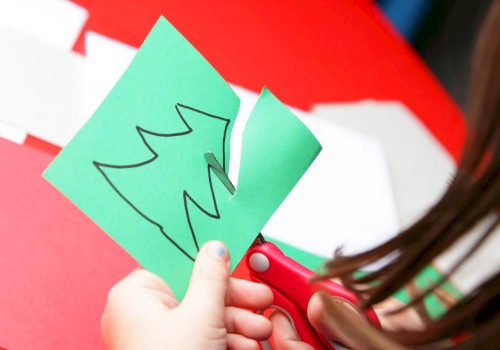 A person is cutting out the shape of a Christmas tree from a green piece of paper with scissors.