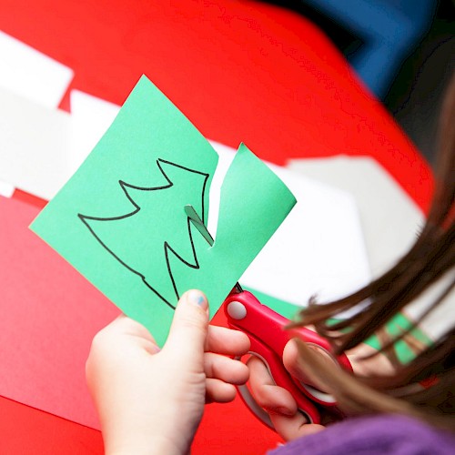 A person is cutting out the shape of a Christmas tree from a green piece of paper with scissors.