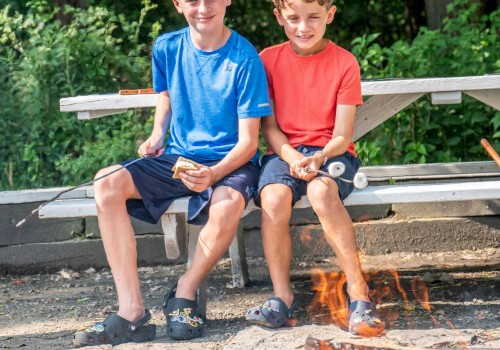 Two boys are sitting on a park bench, roasting marshmallows over a campfire in a lush, green outdoor setting.
