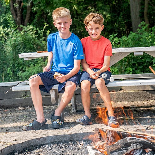 Two boys are sitting on a park bench, roasting marshmallows over a campfire in a lush, green outdoor setting.