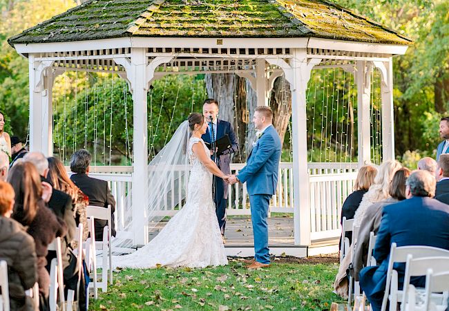 A couple is getting married outdoors under a gazebo surrounded by guests seated in white chairs and bridal party members standing nearby.