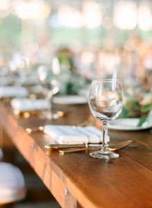 A long wooden table set elegantly with glassware, white plates, gold utensils, napkins, and green floral arrangements in a well-lit venue.