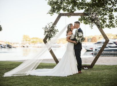 A couple in wedding attire stands under a hexagonal wooden arch adorned with flowers, next to a waterfront with docked boats.