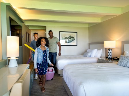 A family of three is entering a hotel room with two beds, carrying luggage and a pool noodle, while smiling and looking happy.