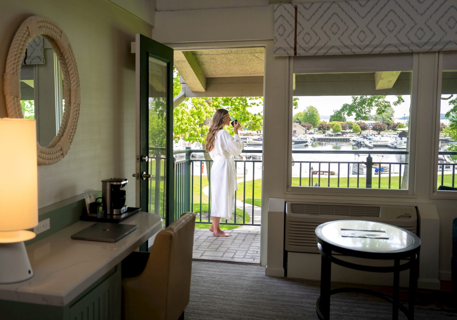 A person in a bathrobe is standing on a balcony, looking at the view. The room has a desk, lamp, and coffee maker. Sunlight brightens the space.