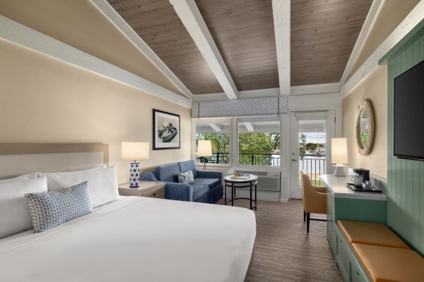 A cozy hotel room with a bed, sofa, TV, and desk. Art on the wall, a coffee maker on the green cabinet, and a window with a scenic view.