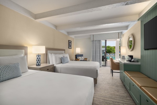 The image shows a hotel room with two beds, a TV, a desk, a mirror, and a view of a balcony, decorated in neutral tones.