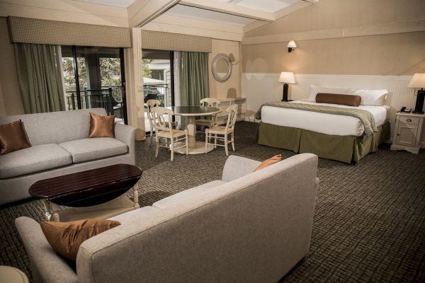 A spacious hotel room features a king-sized bed, a sitting area with sofas, a coffee table, a dining set, and large windows with drapes.