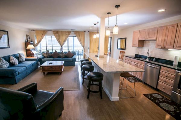 A modern open-plan living room and kitchen with wooden floors, a blue sofa, armchairs, a coffee table, and a granite kitchen island with stools.