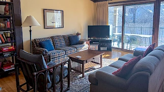 A cozy living room with a plaid sofa, armchairs, coffee table, bookshelf, floor lamp, TV on a stand, and a sliding glass door leading to a balcony.