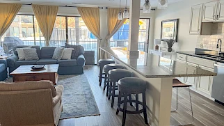A modern open-plan living area with a gray sofa, beige chairs, and a wooden coffee table, plus a bright white kitchen island and bar stools, large windows with curtains.