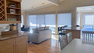 A bright, open living area with light wood floors, beige sofas, a kitchen island, and plenty of windows with blinds letting in soft daylight.