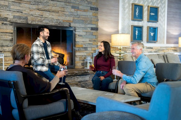 A group of four adults sits in a modern living room, chatting and toasting with drinks by a fireplace and cozy seating.