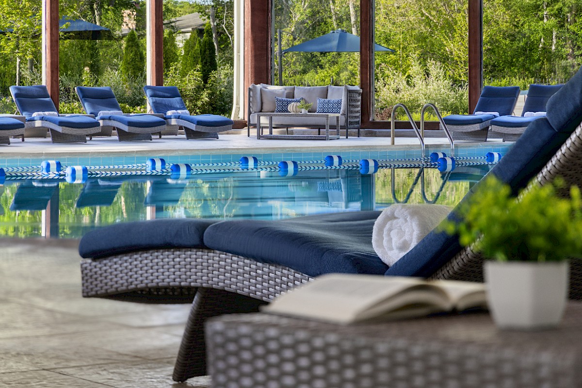 A luxurious outdoor pool area with blue lounge chairs, a calm pool, shaded seating, and a book on a wicker table, all under sunny skies.