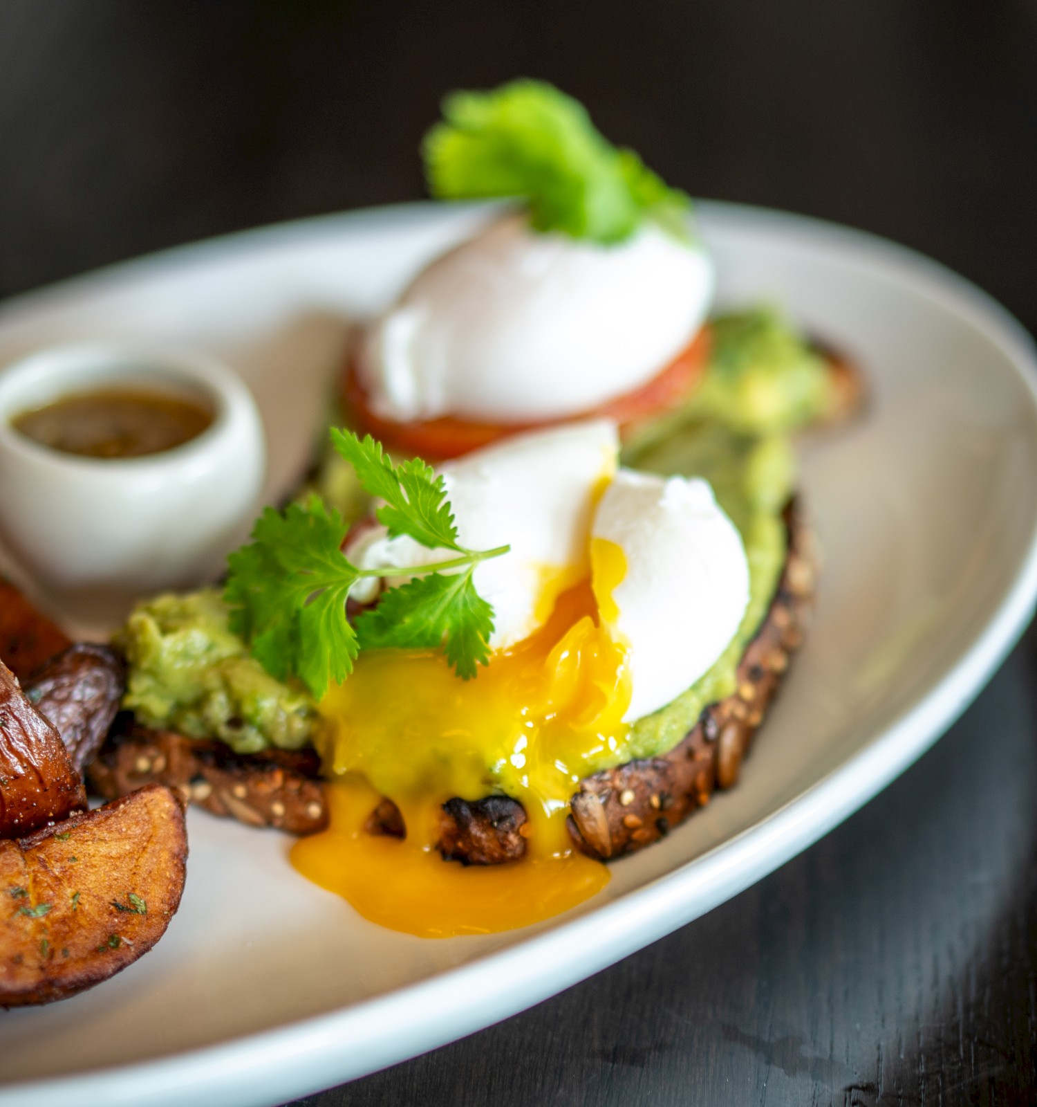 The image shows poached eggs on toast with avocado and a side of roasted potatoes, garnished with herbs, served on a white plate.