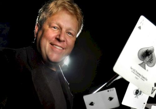 A person is smiling while holding playing cards, including the ace of spades, in a dynamic pose against a dark background.