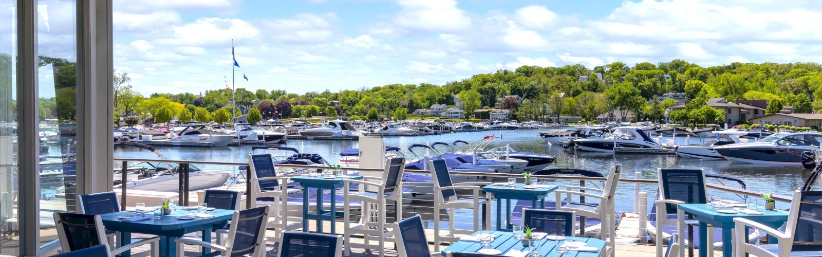 Outdoor dining area with blue tables and chairs overlooks a marina filled with boats under a partly cloudy sky.