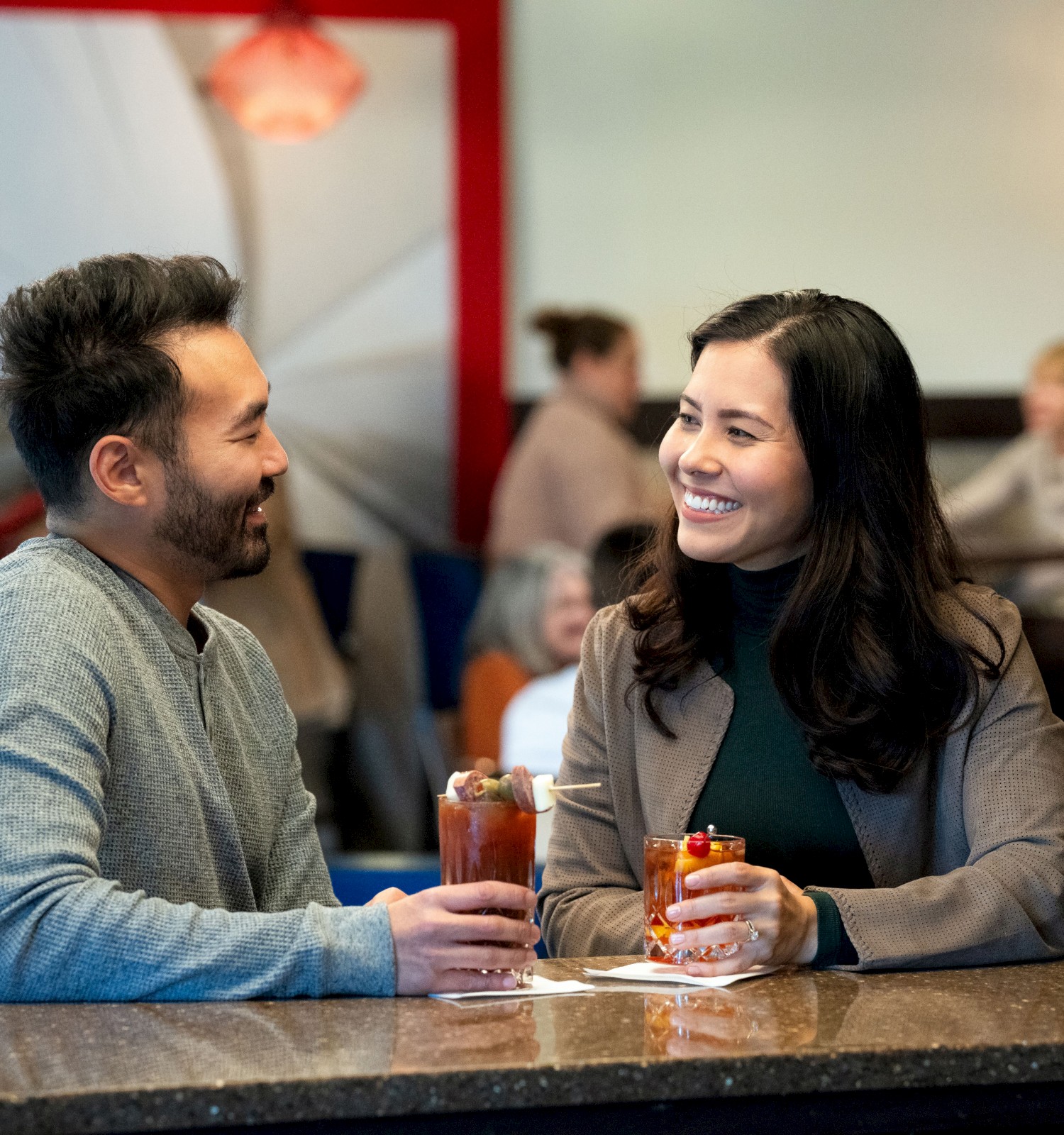 A man and woman are smiling at each other while holding drinks at a bar, with others in the background.