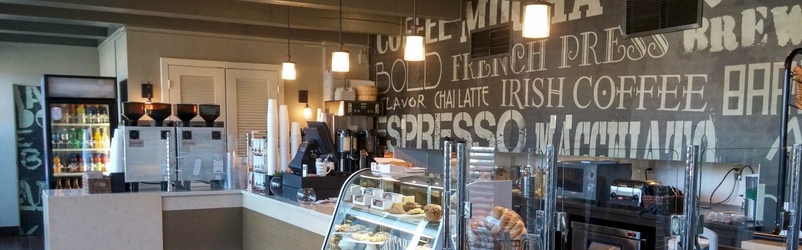 A modern coffee shop with a pastry display, beverage dispensers, refrigerated drinks, and decor featuring coffee-related words. Customers are absent.