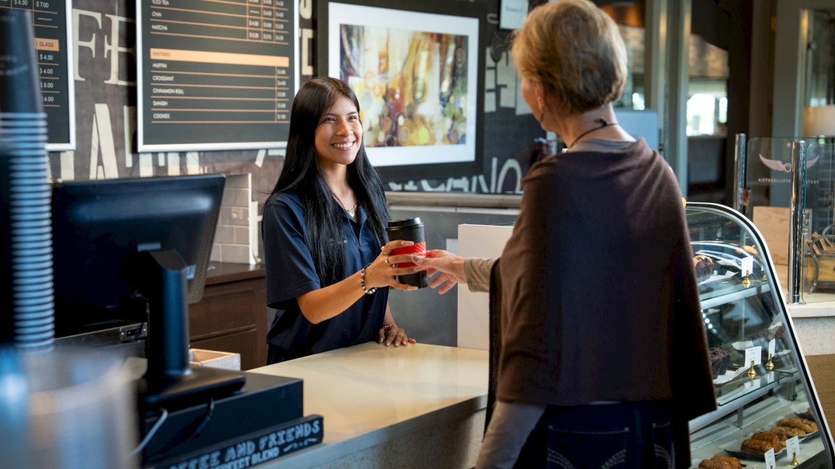 A customer receives a drink from a cashier at a café. A display case with pastries is nearby, and a menu is visible in the background.