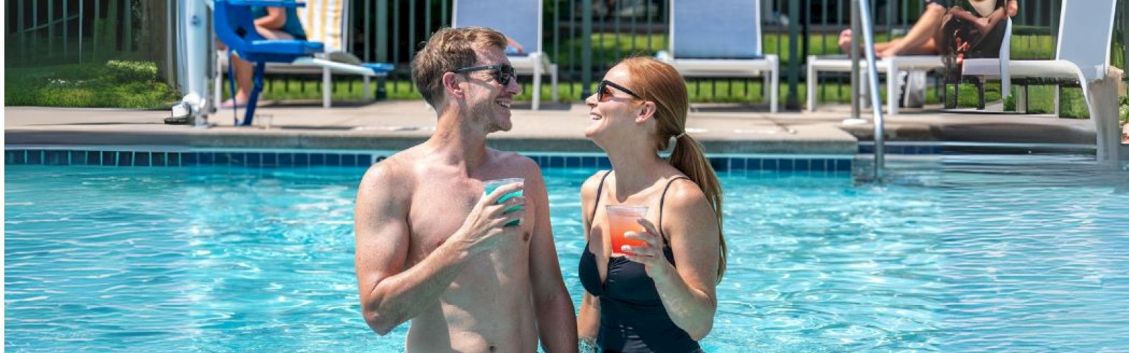 A man and woman enjoy drinks while standing in a swimming pool, with others relaxing by the poolside on lounge chairs.