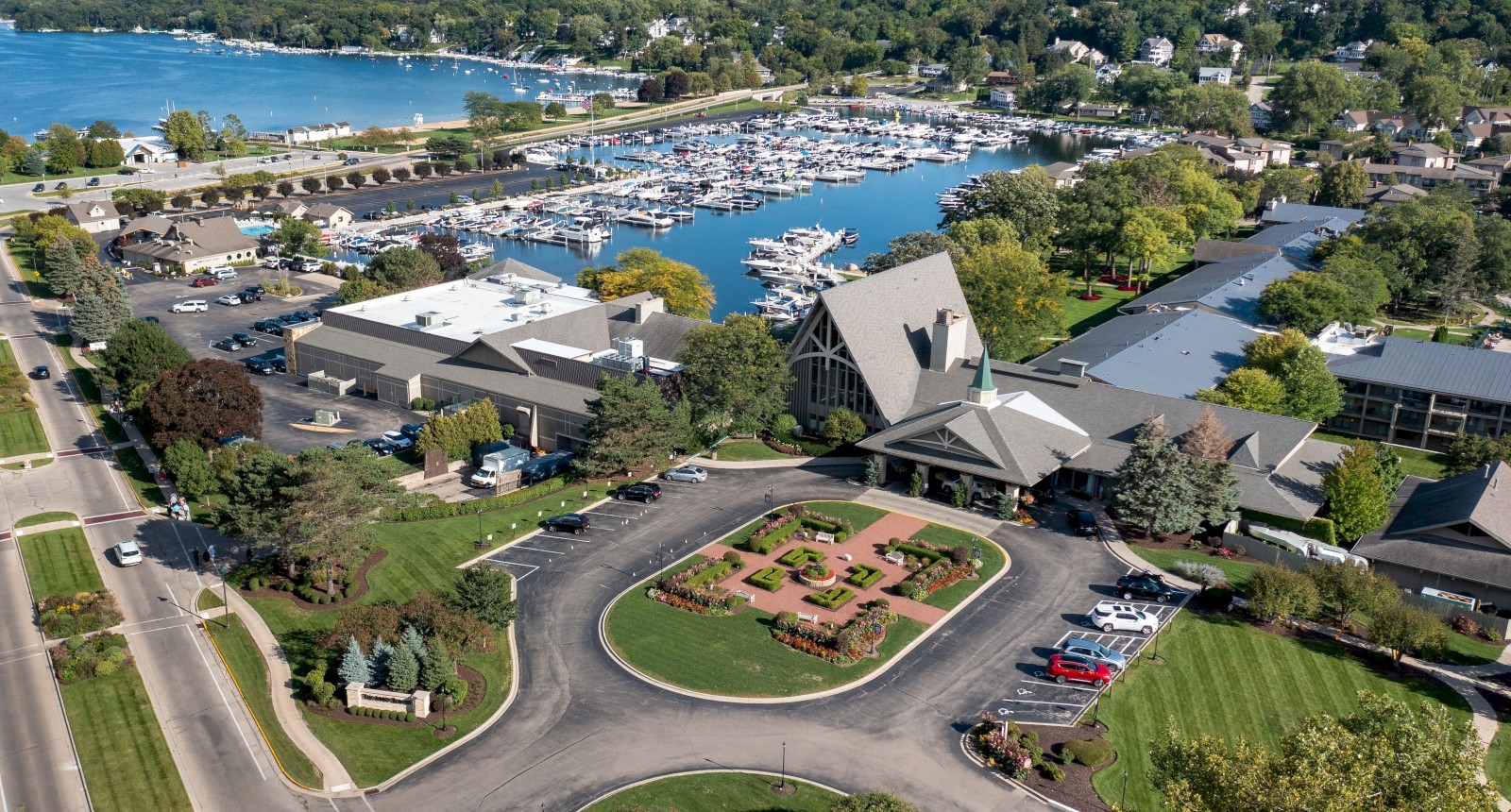 An aerial view of a lakeside marina with numerous boats, surrounded by greenery, buildings, roads, and landscaped areas with pathways and cars.