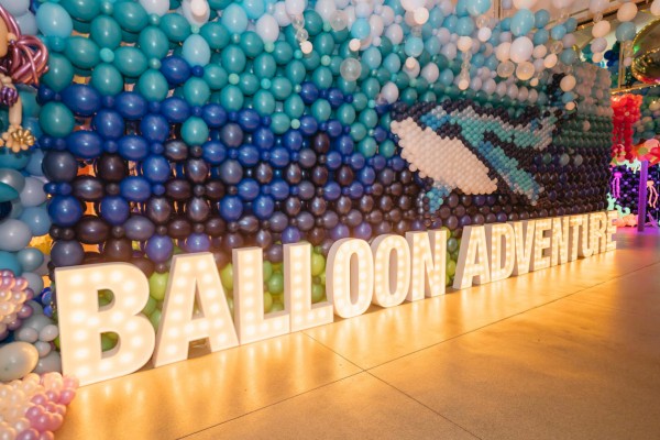 A wall covered in teal, blue, and white balloons forming a gradient, with large illuminated letters spelling “BALLOON ADVENTURE” along the base.