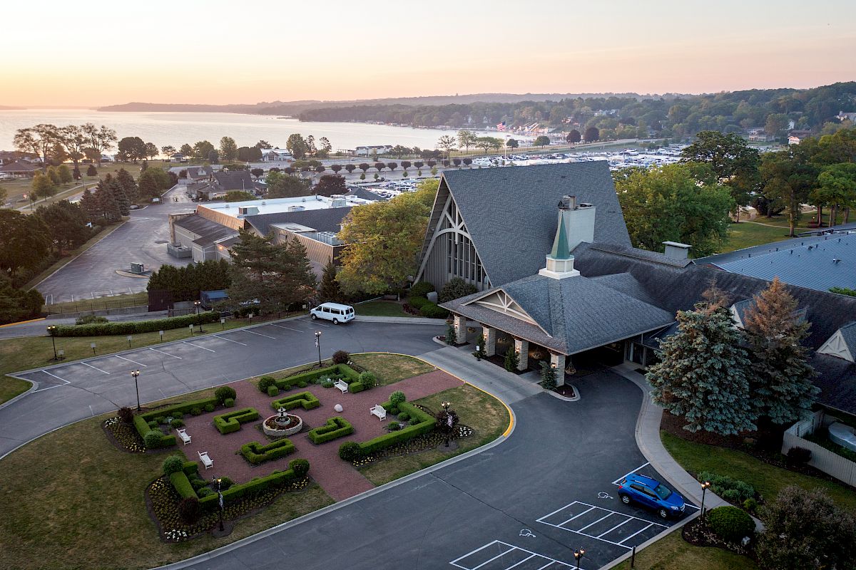 An aerial view of a building with a landscaped garden, near a body of water and a parking area, surrounded by trees and additional structures.