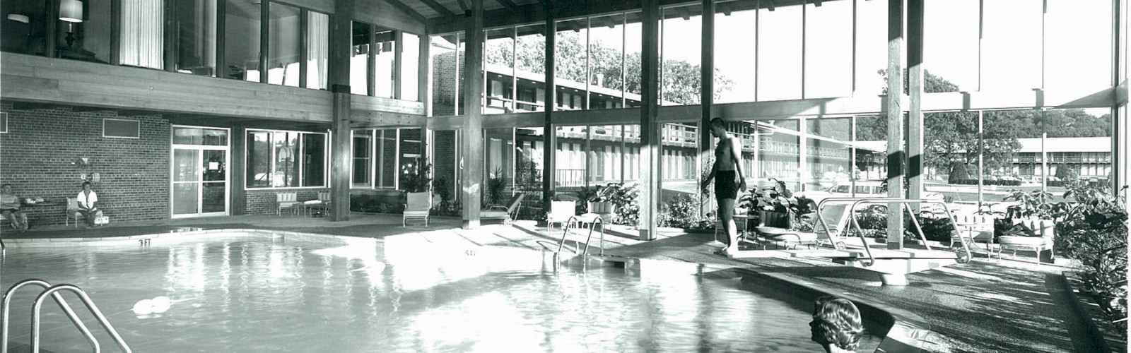 A large indoor pool area with high wooden ceilings, glass walls, and several people around the pool, including one seated on the pool's edge.