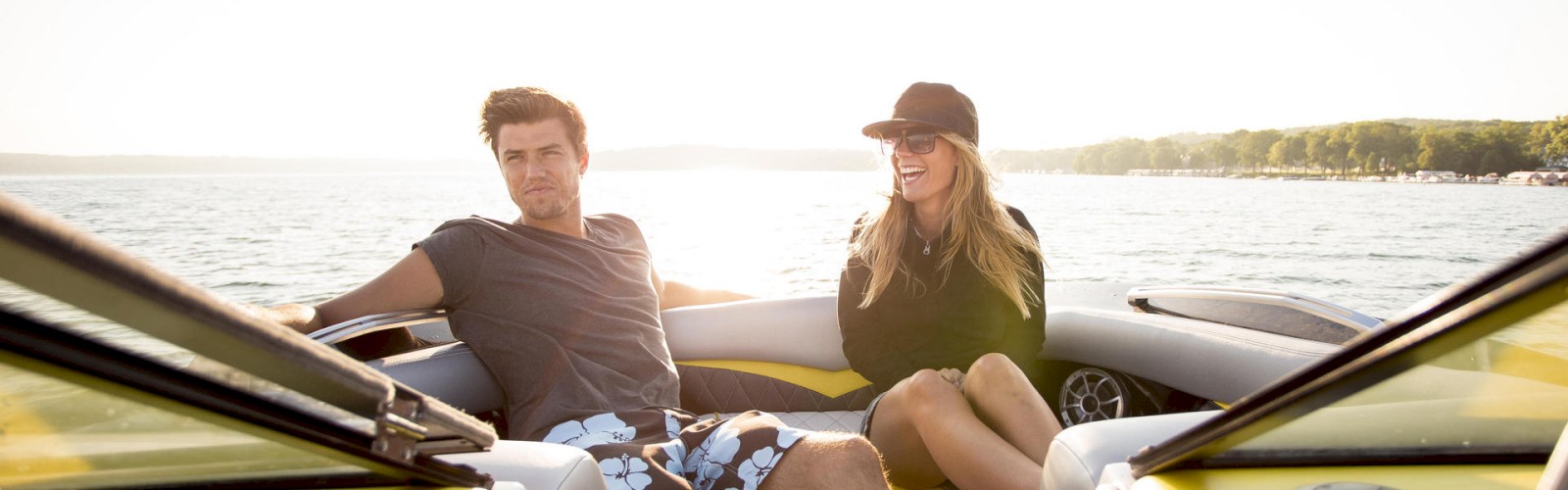Two people enjoying a sunny day on a boat in casual attire, sitting comfortably near the front, with water and distant greenery in the background.