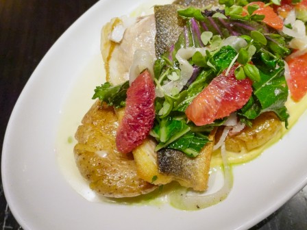 A plate of seared fish with citrus sauce, greens, and colorful citrus segments on top.