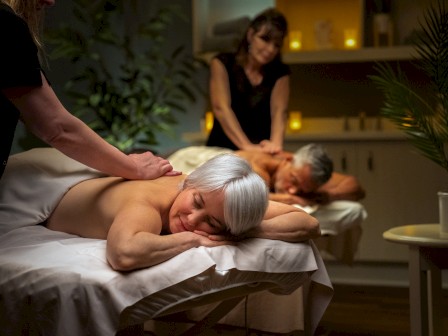 Two individuals are receiving a massage in a serene, dimly-lit spa setting with soft lighting and candles, creating a relaxing ambiance.
