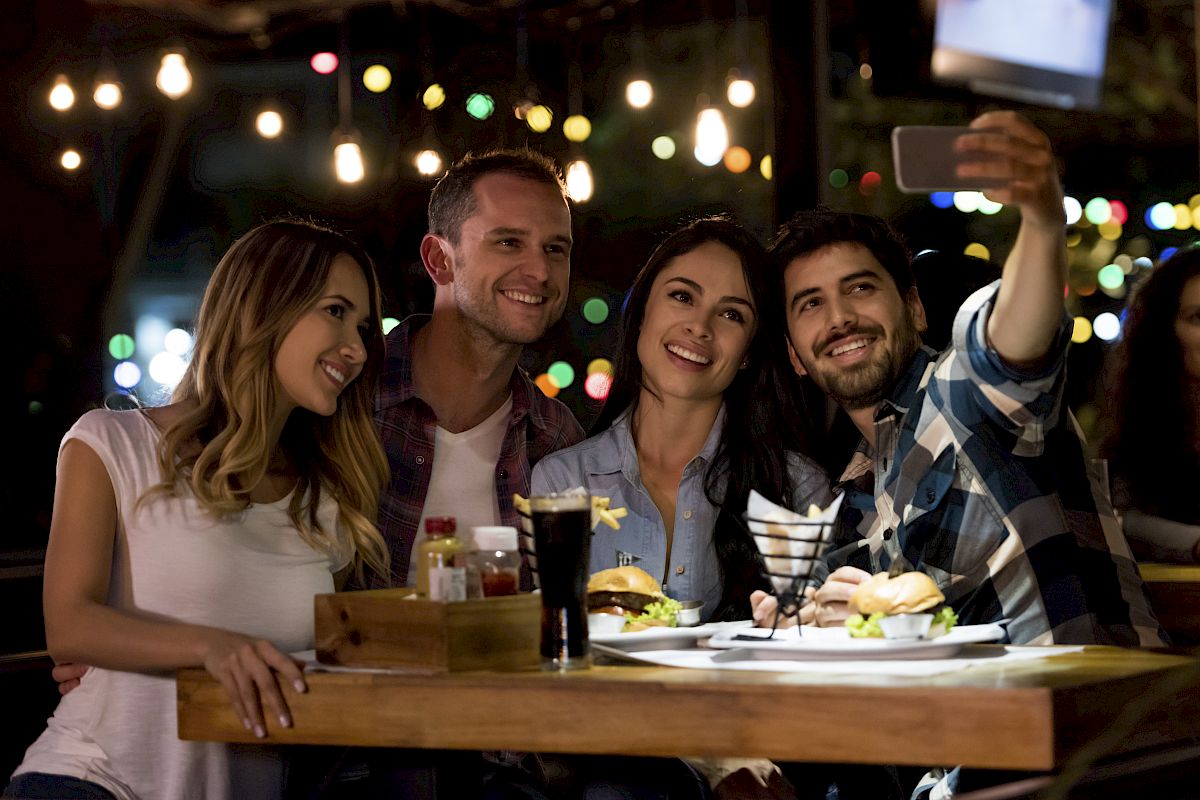 A group of four friends at a restaurant are smiling and taking a selfie together at a table with burgers, drinks, and hanging lights in the background.