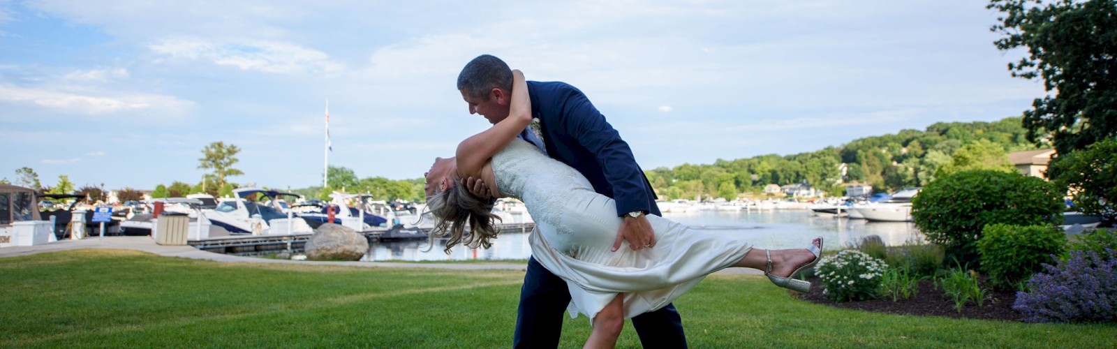 A couple is dancing on a grassy area near a marina, with boats and greenery in the background.