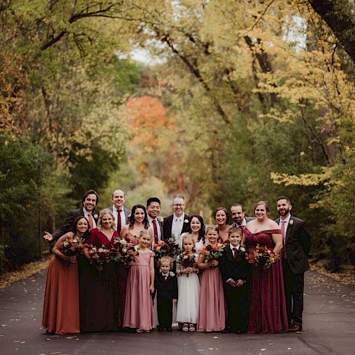 A wedding party poses outdoors on a tree-lined road with colorful autumn leaves, people smiling and dressed in formal attire, holding flowers.