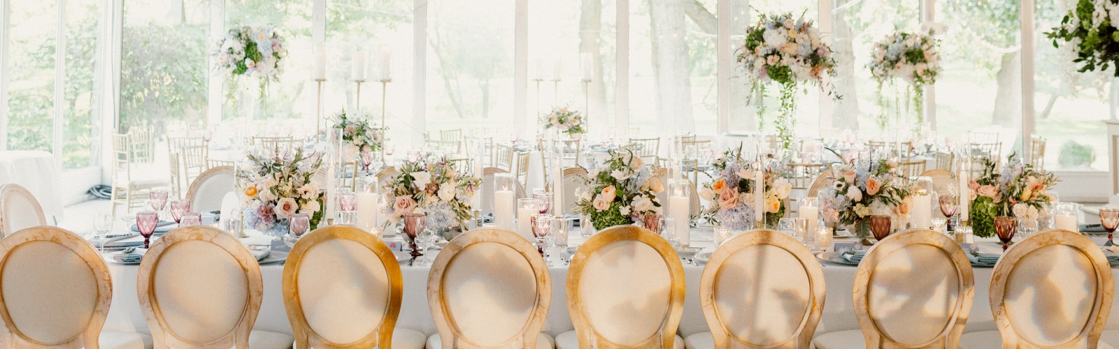 An elegant banquet setup under a white canopy with chandeliers, round-backed chairs, floral centerpieces, and table settings ready for guests.