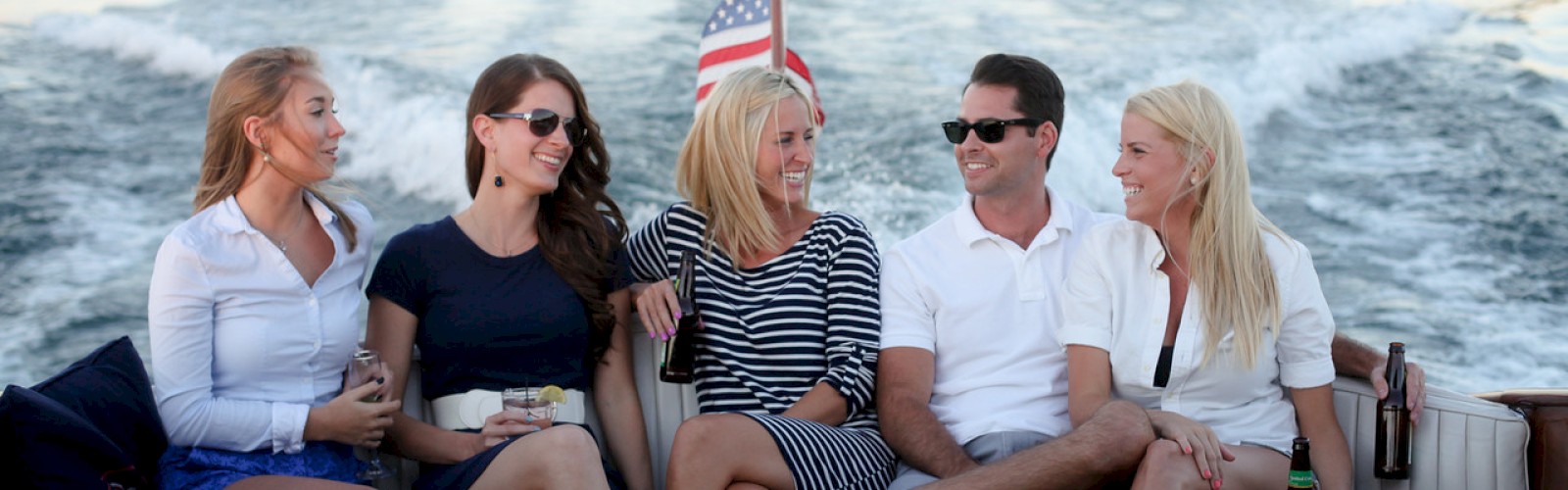 Five people sitting on a boat, smiling and talking, with an American flag and a scenic water view in the background.