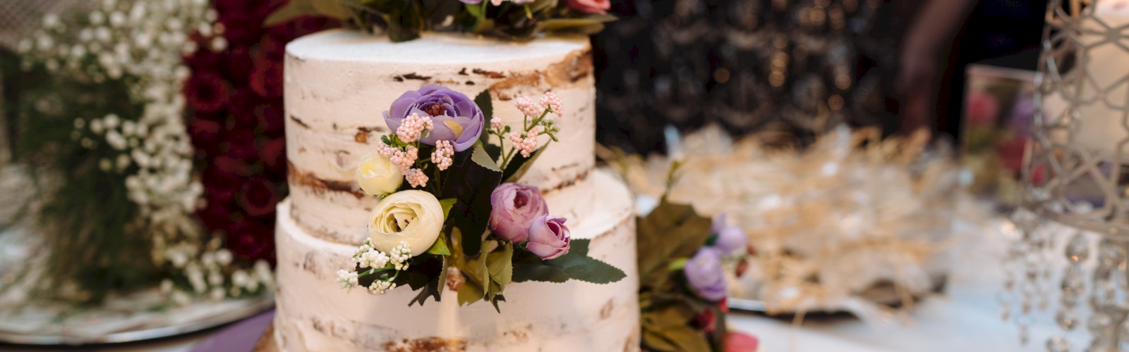 A two-tiered cake decorated with flowers sits on a wooden base. There are floral arrangements and a candle holder in the background.