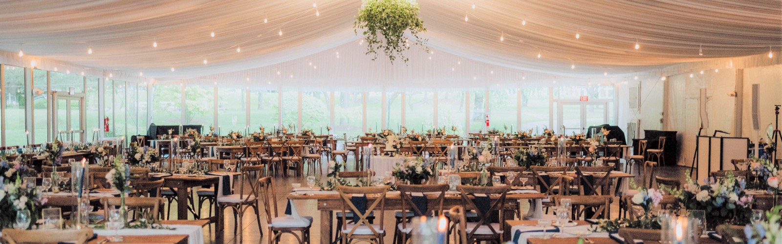 A beautifully decorated venue with draped ceilings, wooden chairs, tables set for a formal event, and hanging greenery under soft lighting.