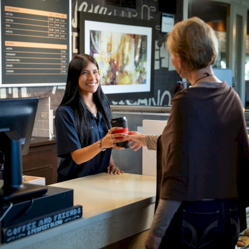 A customer receives a drink order from a smiling barista at a cozy coffee shop counter.
