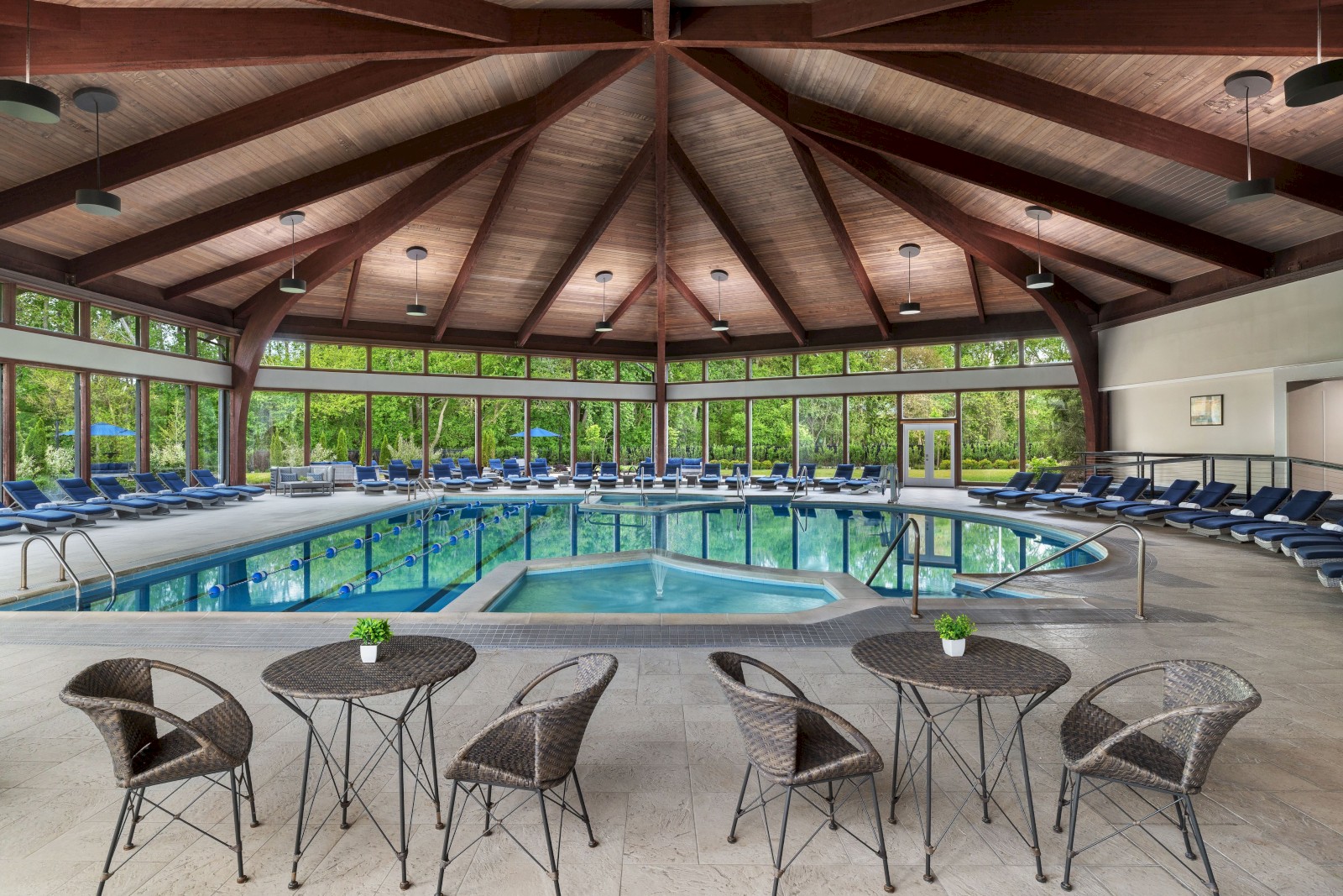 An indoor pool area with a high wooden ceiling, surrounded by lounge chairs, tables, and chairs, with large windows showing greenery outside.