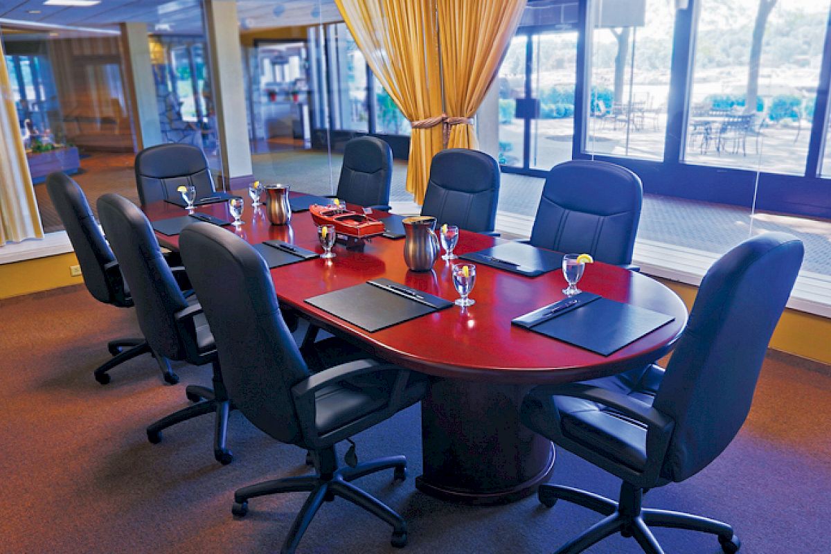 The image shows a conference room with a wooden table, eight leather chairs, water glasses, notepads, and windows with a view of an outdoor area.