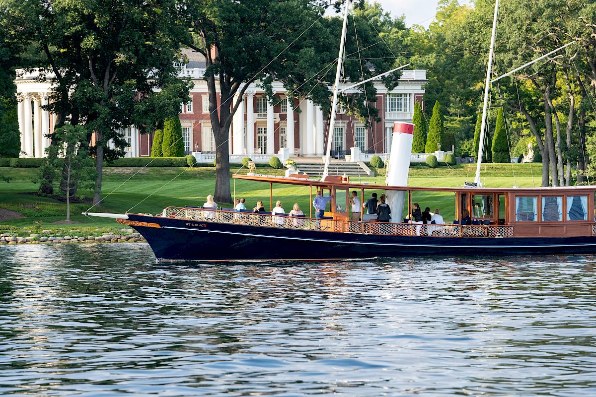 A boat sails on a lake with a group of people on it, passing by a grand house with pillars surrounded by lush trees and manicured lawns.