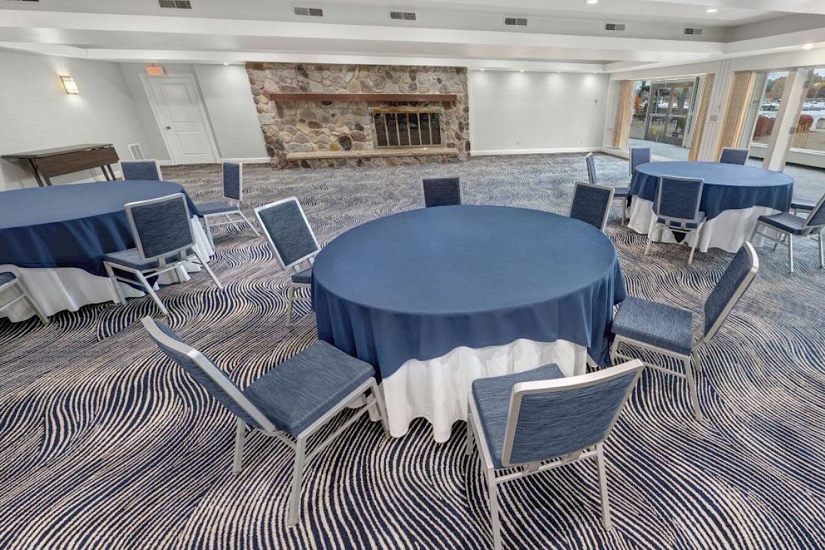 The image shows an empty conference room with round tables, blue tablecloths, chairs, a patterned carpet, large windows, and a stone fireplace.
