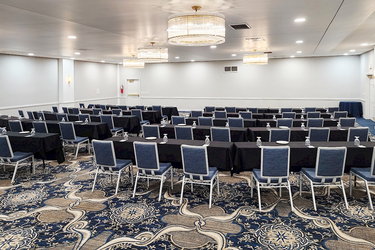 A conference room is set up with rows of blue chairs and tables with water bottles, under chandeliers on a patterned carpet.