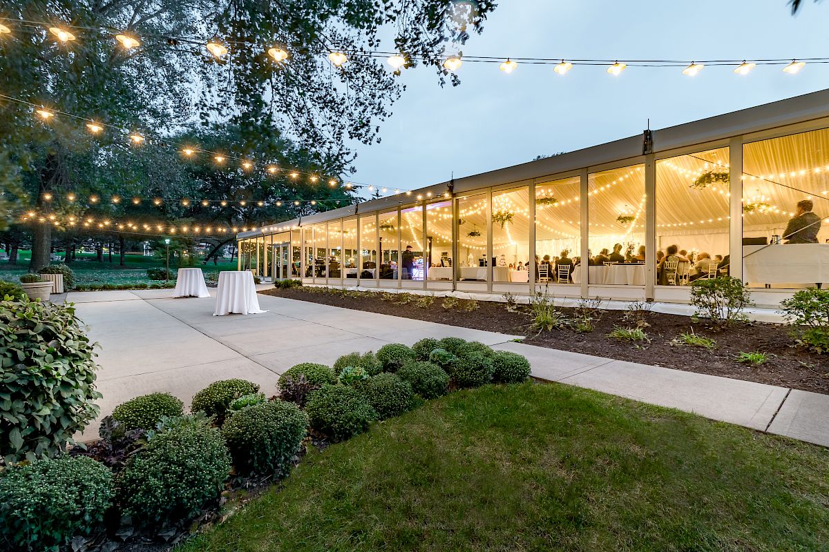 An outdoor event venue with string lights and a tent canopy is seen, with tables inside and well-manicured greenery surrounding the area.