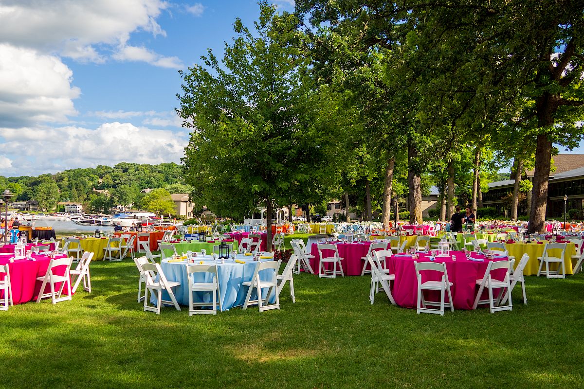 An outdoor event setup with round tables covered in colorful tablecloths and white chairs on a grassy area near a waterfront, under a bright sky.