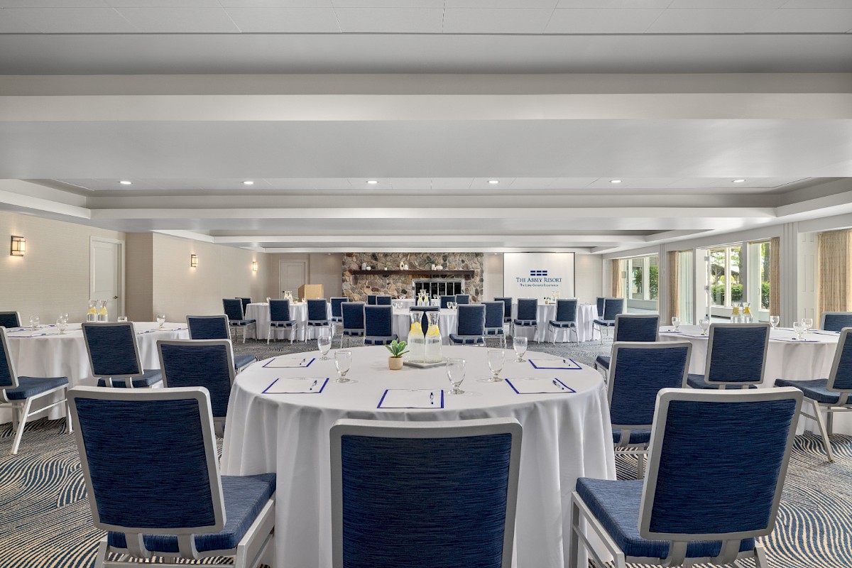 A conference room with round tables and blue chairs is set up for an event, with place settings and a presentation screen at the front.